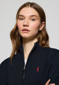 Pull à zip bleu marine fabriqué en tissu tricoté, avec un col haut et un logo rouge sur la poitrine gauche. Texture lisse et design ajusté.