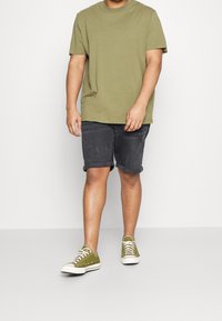 Olijfgroene t-shirt met korte mouwen, gecombineerd met donkergrijze denim shorts met opgerolde pijpen. Schoeisel bestaat uit groene low-top sneakers met witte zolen.