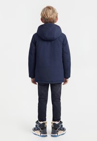 Lyshåret barn iført en marineblå hættetrøje, mørke bukser og blå sneakers står med ryggen til på en ensfarvet baggrund.