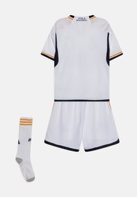 Conjunto de camiseta y pantalones cortos deportivos blancos con detalles en marino y amarillo, que presenta el texto “¡HALA MADRID!” en el cuello. Un calcetín blanco con rayas.