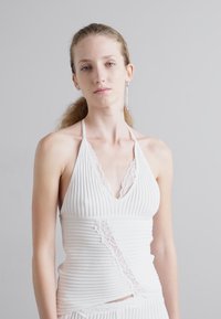 Haut halter blanc à côtes avec des accents en dentelle, présentant un design en V et un motif asymétrique, fabriqué en tissu texturé.