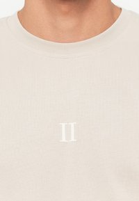 Light beige crewneck t-shirt made of soft fabric, featuring a small white "II" logo on the front, with a smooth texture and ribbed neckline.
