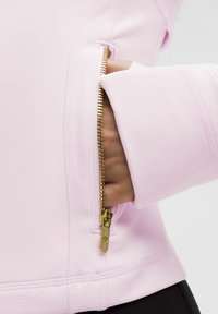 Jasnoróżowa kurtka z złotym zamkiem, z ręką częściowo wsuniętą w kieszeń na zamek. Miękka faktura i dopasowany krój.