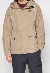 Beige lätt jacka med huva, två framfickor, elastiska ärmmuddar och en texturerad yta. Har en liten svart logotyp på bröstet.