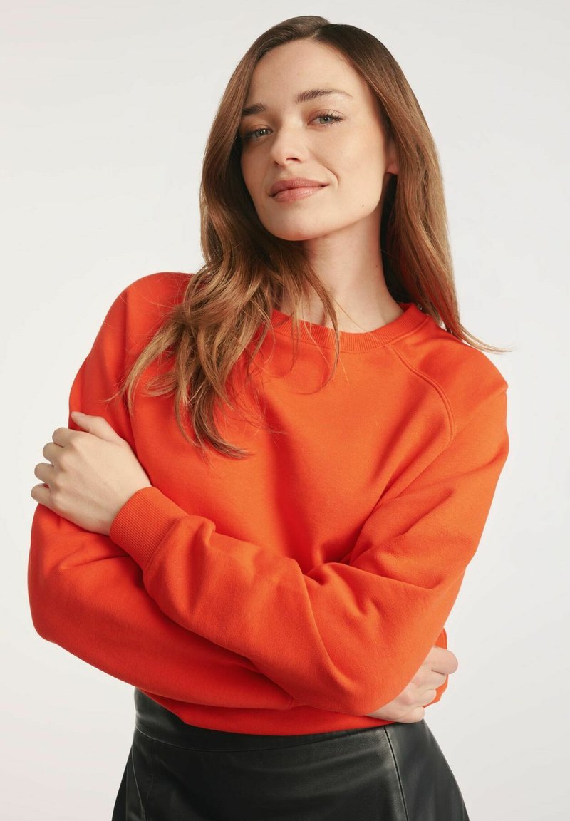 Jasnopomarańczowy sweatshirt z okrągłym dekoltem i długimi rękawami, wykonany z miękkiego materiału. Modelka nosi czarną spódnicę.