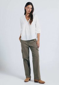 White long-sleeve blouse with pleated front paired with olive green cargo pants. Model wears tan ankle boots and layered necklaces.