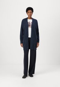 Cardigan albastru marin, cu o textură moale, cu deschidere în față și fără închizători, purtat peste o cămașă albă cu un imprimeu grafic, asortat cu pantaloni închiși la culoare și încălțăminte.