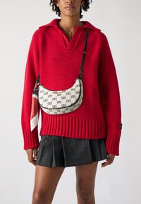 Strikket rød sweater med bred krave, kombineret med en plisseret sort nederdel. En lille cremefarvet taske med et logo mønster og stofscarf som accent.