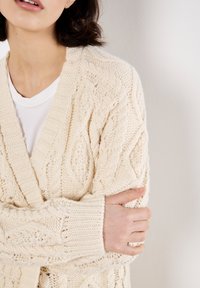 Cremfarbener Strickcardigan mit strukturiertem Zopfmuster, offener Vorderseite und gerippten Bündchen. Der Stoff wirkt dick und warm.