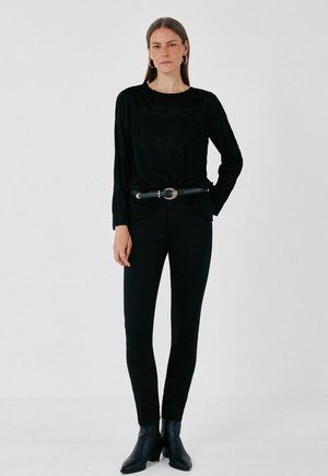 Femme debout portant un haut noir à manches longues, un pantalon noir slim avec ceinture, des bottines noires et des boucles d'oreilles créoles dorées sur un fond uni.