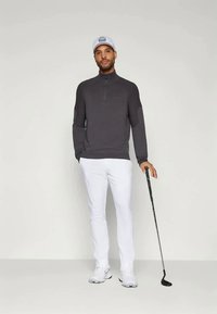 Männliches Golfoutfit mit einem dunkelgrauen Reißverschluss-Pullover und weißen Hosen, kombiniert mit einer weißen Kappe und einem Golfschläger. Weicher Stoff, enganliegendes Design.