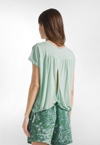 Mintgrøn bluse med løs pasform, korte ærmer og en nøglehuldetalje bagpå. Kombineret med grønne shorts med bladmønster.