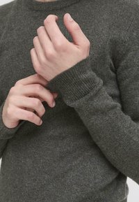 Pulover tricotat de culoare gri închis, cu guler rotund. Mâneci lungi cu manșete ribbate. Materialul pare moale, cu o ușoară textură. Mâini care ajustează mâneca.