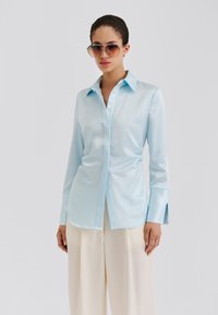 Φωτεινό γαλάζιο σατέν πουκάμισο με γιακά με κουμπιά, μακριά μανίκια και πλαϊνές σκισμές. Συνδυασμένο με κρεμ φαρδιά παντελόνια.