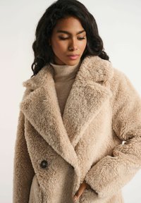 Beżowy, dwurzędowy płaszcz z puszystej, teksturowanej tkaniny; ma szeroki kołnierz i zapięcie na guziki. Stylizowany na beżowym golfie.