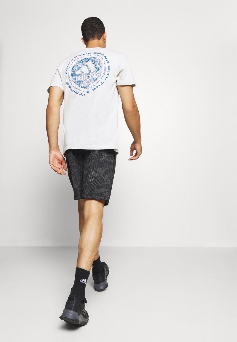 Man som går bort iklädd en vit Adidas T-shirt med blå logotyp, svarta mönstrade shorts, svarta strumpor och svarta sneakers på en neutral bakgrund.