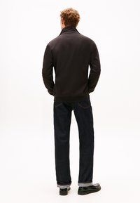 Mand med krøllet hår iført en sort jakke, mørkeblå op rullede jeans og sorte loafers, stående med ryggen til på en hvid baggrund.