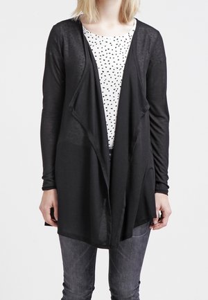 Black long-sleeve cardigan with an open front and draped design, worn over a white shirt with small black triangles, paired with gray jeans.