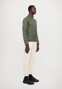Green puffer jacket with horizontal stitching, high collar, and zip closure. Paired with light-colored sweatpants and black sneakers.