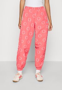 Donna che indossa pantaloni da jogging Adidas di colore corallo con loghi circolari bianchi, calze bianche e sneakers bianche con righe corallo.