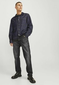 Denim jakna v temno modri barvi z dvema žepoma na prsih, v kombinaciji s črnimi kavbojkami. Model nosi črne čevlje. Gladka tekstura, sproščen kroj.