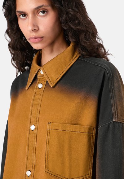 Junge Frau mit dunklem, lockigem Haar, die ein senfgelb-schwarzes Button-Up-Hemd mit einer Brusttasche trägt und leicht zur Seite schaut.