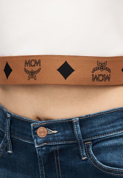 Καφέ λουρί με μαύρες πινελιές που φέρει τα γράμματα «MCM» και ένα φτερωτό έμβλημα, σε συνδυασμό με μπλε τζιν σορτς, με λεπτομέρεια σε χαλκό κουμπί.