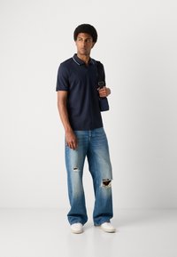 Ναυτικό πόλο μπλουζάκι με γιακά και λευκή πίπα, σε συνδυασμό με ανοιχτό μπλε παντελόνι καμπάνα που έχει μεγάλες τρύπες. Το μοντέλο κρατά μια μικρή τσάντα.