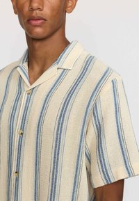 Camisa de manga corta, abotonada, en color crema con rayas verticales azules. Textura tejida con un collar relajado y botones beige.