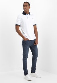 Camisa polo branca com colarinho azul-marinho e detalhes azuis, combinada com calças azul-marinho e ténis brancos. Modelo posa contra um fundo branco.