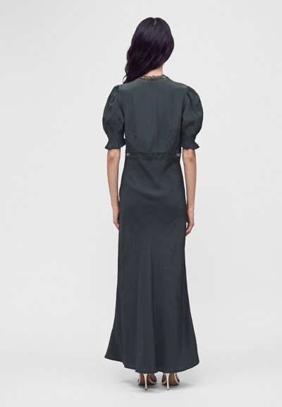 Femme aux longs cheveux foncés portant une longue robe vert foncé à manches courtes bouffantes, debout pieds nus, tournée de dos.