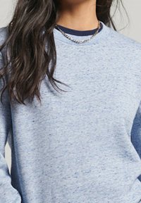 Donna che indossa un maglione a pois blu chiaro sopra una maglietta a girocollo blu navy e una collana d'argento, con lunghi capelli scuri.