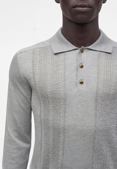 Personne à la peau foncée portant un polo gris clair à manches longues avec des motifs texturés verticaux et trois boutons dorés sur la patte de boutonnage.
