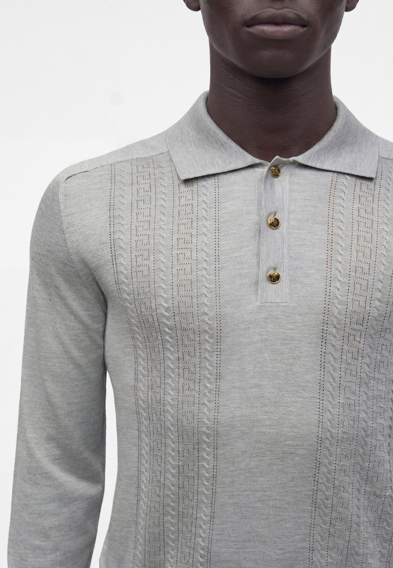 Personne à la peau foncée portant un polo gris clair à manches longues avec des motifs texturés verticaux et trois boutons dorés sur la patte de boutonnage.