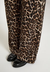 Široké nohavice so vzorom leopardej kože v hnedom a čiernom prevedení, skombinované s lesklými čiernymi mokasínami s špicatou špičkou a dekoratívnym prešívaním.