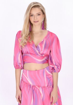 Nainen, jolla on pitkät vaaleat hiukset, pukeutuneena vaaleanpunaiseen ja violettiin satiiniin kietoutuvaan yläosaan, jossa on liehuvat hihat, ja vastaavaan hameeseen, asusteena violetit tupsukorvakorut.