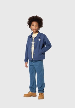 Enfant aux cheveux bouclés portant une veste bleue, un t-shirt graphique beige, un jean bleu et des bottes à lacets couleur beige, debout devant un fond gris uni.