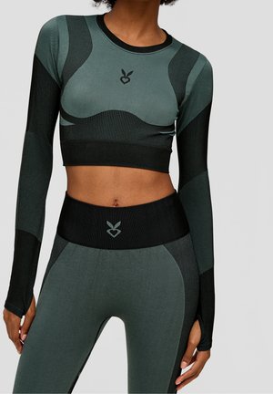 Kvinde iført matchende grøn og sort langærmet crop top og højtaljede leggings med lille hjerte- og bladlogo på bryst og talje.