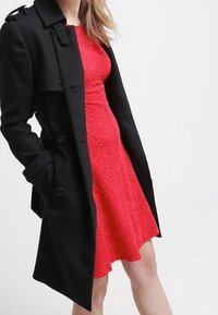 Svart trench coat över en strukturerad röd klänning. Kappan har en dubbelknäppt front och axelremmar, vilket skapar en lagerlook.