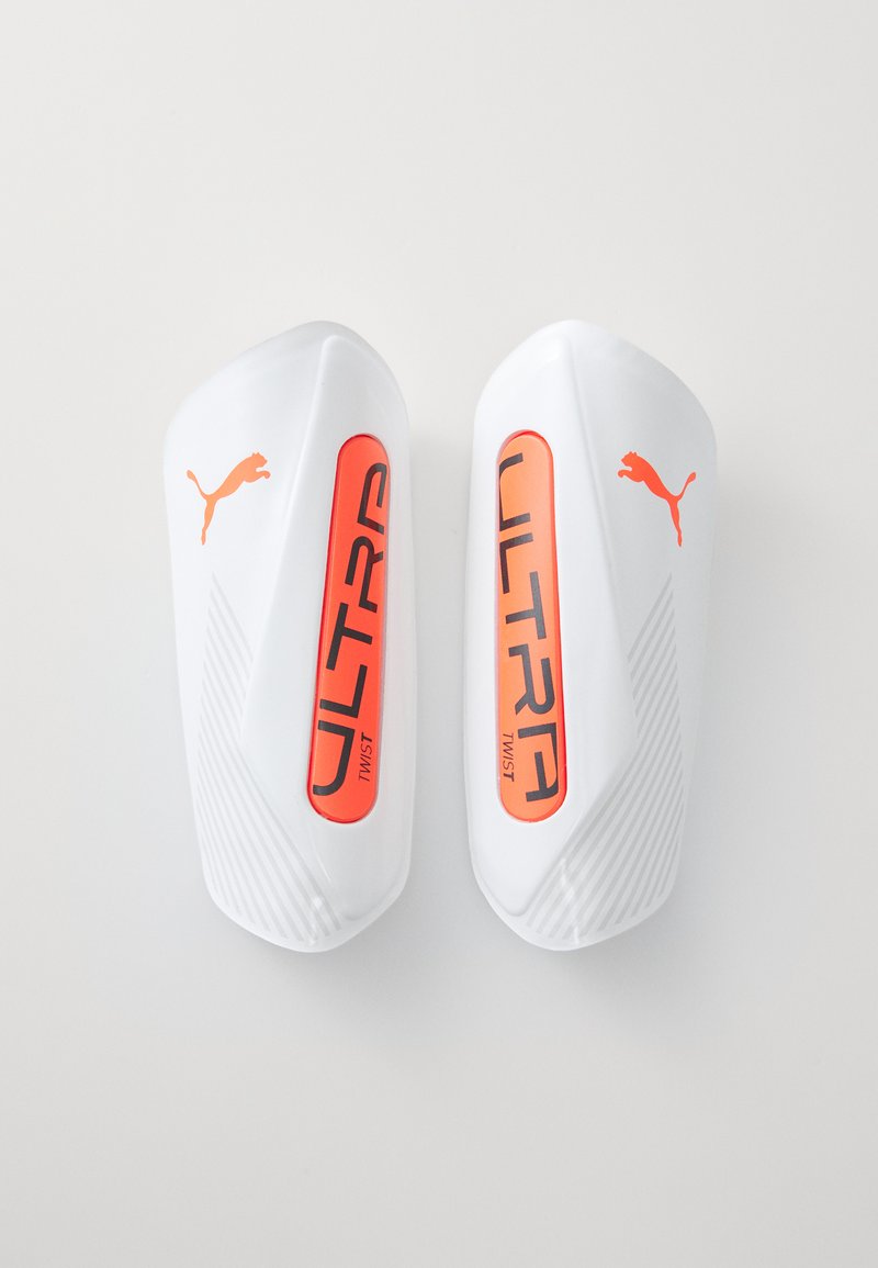 Puma ULTRA chrániče holeních v bílé plastové barvě s oranžovými akcenty. Vyznačují se texturovaným designem a výrazným brandingem "ULTRA TWIST".