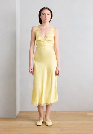 Kobieta stojąca w bezrękawowej, jasnożółtej sukience midi z satyny z koronkowym wykończeniem i pasującymi wsuwanymi butami na tle jednolitej ściany.