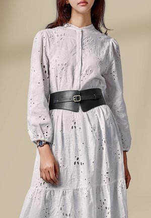 Frau trägt ein weißes Blumenspitzenkleid mit gerafften Ärmeln und einem schwarzen, breiten Gürtel mit Schnalle, beiger Hintergrund.