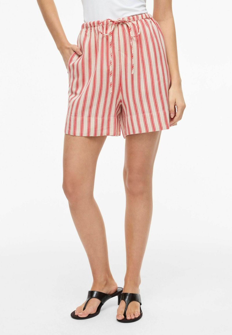 Femme portant un short beige et rouge à rayures verticales avec cordon de serrage et poches, associé à des sandales à talons noires à bouts ouverts.