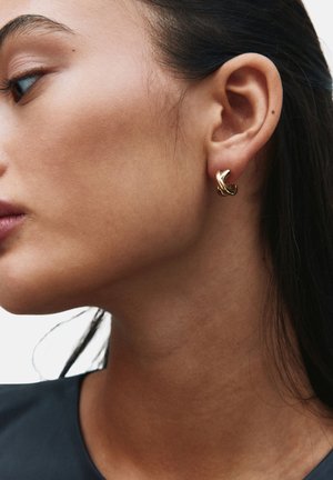 Kvinde med mørkt hår iført en snoet guldring i venstre øre, vist i nærbillede profil mod en lys baggrund.