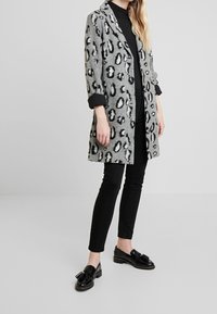 Grijze panterprintjas met zwarte accenten, lange mouwen, open voorkant, gecombineerd met zwarte skinny broeken en zwarte loafers met franjes.