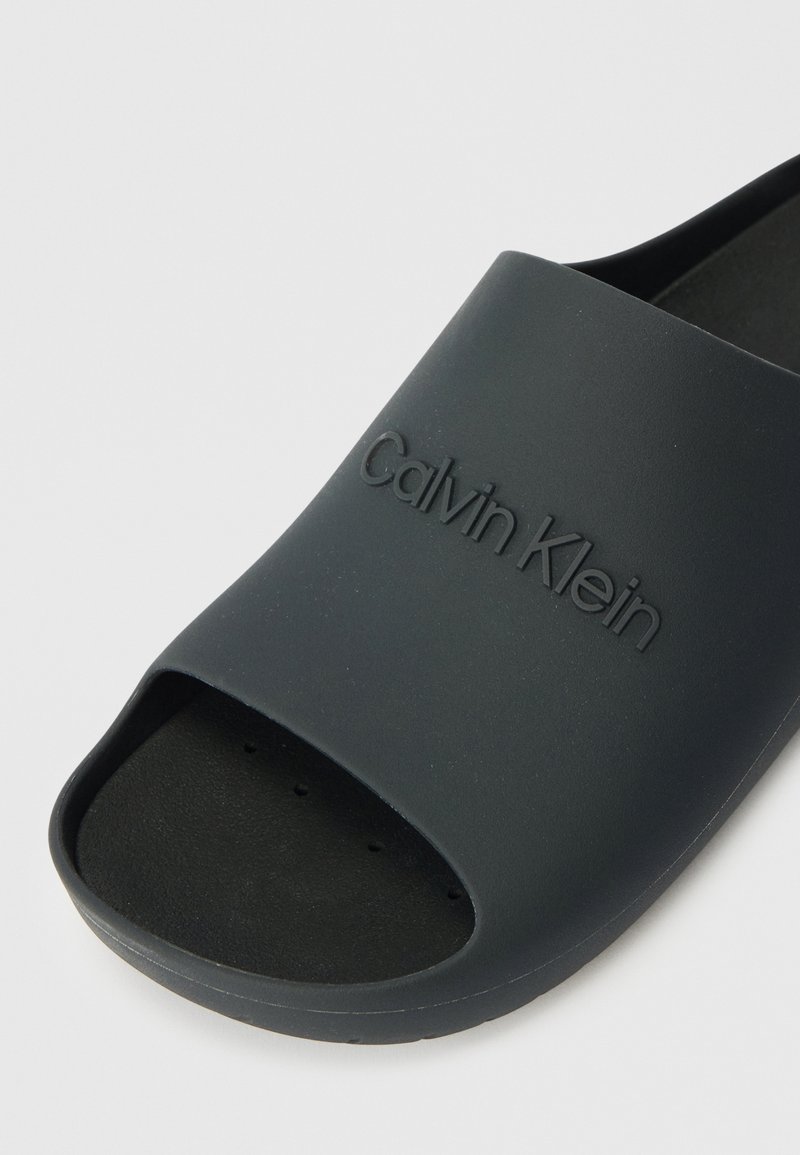 Schwarze Gummisandale mit offenem Zeh, strukturiertem Fußbett und geprägt "Calvin Klein"-Logo auf dem Riemen. Glatte Oberfläche.