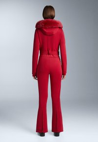 Piros síruha műszőrme gallérral, testre szabott szabás, derékon övvel, és bővülő szárú nadrággal, az alján cipzárokkal. Sima textúrájú.
