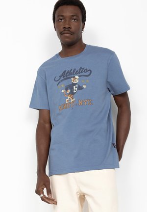Mann trägt ein blaues T-Shirt mit einem Cartoon-Tiger in einem Sporttrikot und dem Text „Athletic Schott. NYC“ in einem Studio.