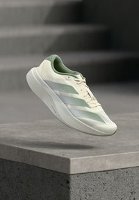 Λευκό και πράσινο παπούτσι για τρέξιμο Adidas με τρεις ρίγες, αιωρούμενο πάνω από μια τσιμεντένια άκρη και σκάλες στο φόντο.