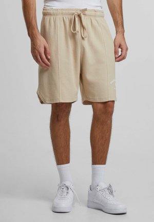 Beige textured shorts with an elastic waistband and drawstring, featuring side slits and a small logo on the left leg.